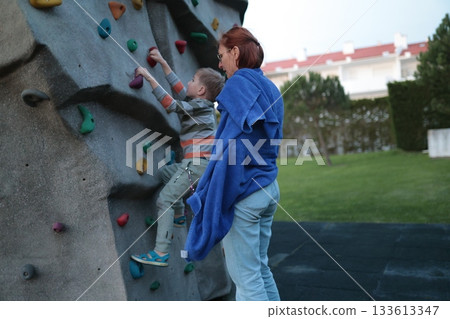 Young boy climbing rock wall with adult support 133613347