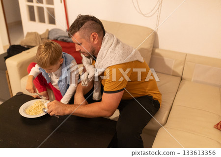 Father helping young son eating pasta at home 133613356