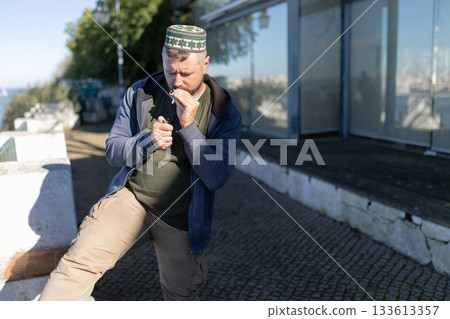 Man in kufi lighting cigarette outdoors during day Man in kufi lighting cigarette outdoors during day 133613357