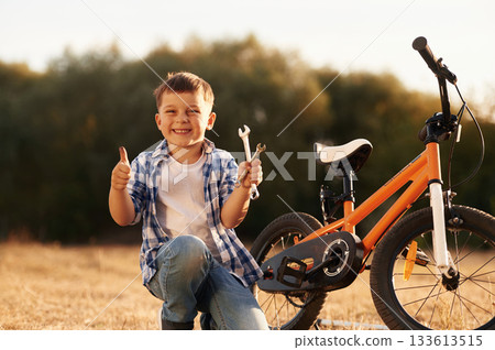 Repairing the bike, holding wrench. Cute boy is with his bicycle on the field Repairing the bike, holding wrench. Cute boy is with his bicycle on the field 133613515