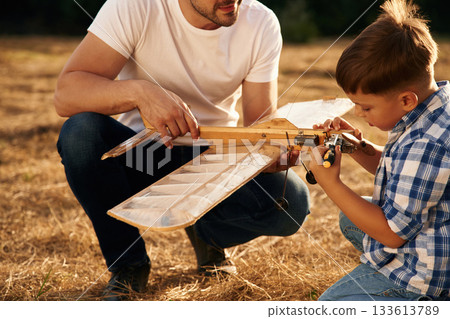 Messing around with broken toy. Father and son are with wooden plane on the field Messing around with broken toy. Father and son are with wooden plane on the field 133613789