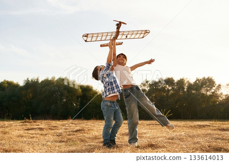 Flying high up. Boy and girl are playing with wooden toy plane on the field Flying high up. Boy and girl are playing with wooden toy plane on the field 133614013