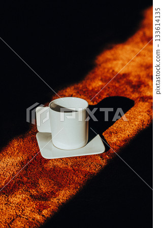 A white coffee cup on a saucer casts a long shadow across a textured brown soft carpet, contrasting with the bright light. Morning drink at cozy home. A white coffee cup on a saucer casts a long shadow across a textured brown soft carpet, contrasting with the bright light. Morning drink at cozy home. 133614135