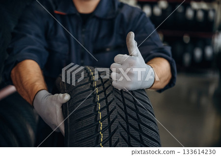 Focused close up view, thumb up, holding a tire. Man worker is maintenance station with wheels 133614230