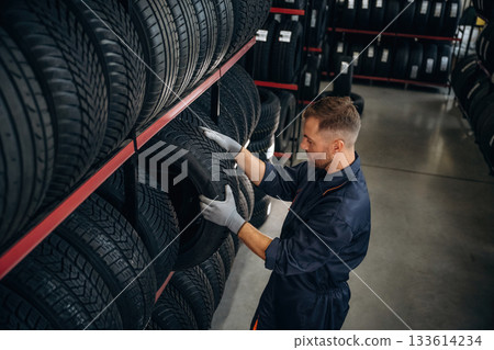 In professional uniform near the shelve. Man worker is maintenance station with tires, wheels In professional uniform near the shelve. Man worker is maintenance station with tires, wheels 133614234