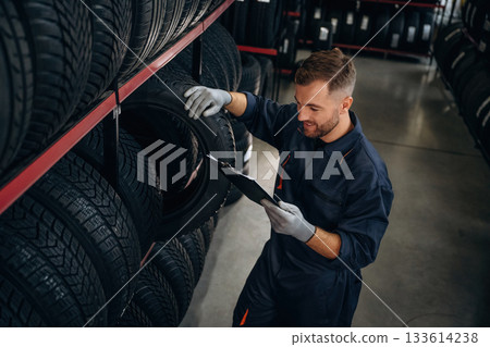 In professional uniform near the shelve. Man worker is maintenance station with tires, wheels 133614238