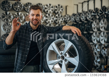 Smiling, positive facial expression, hand gesture. Man worker is maintenance station with tires, wheels 133614388