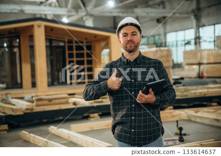 Digital tablet is in hand. Male builder is at construction of wooden house 133614637