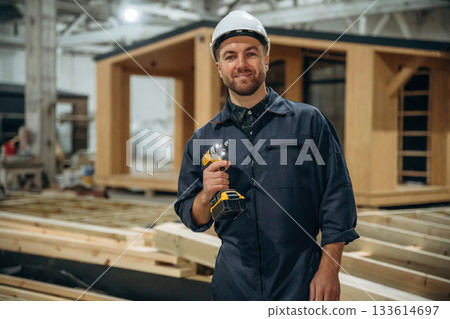 Small drill in hands. Male builder is at construction of wooden house 133614697