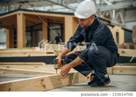 Small drill in hands. Male builder is at construction of wooden house 133614700