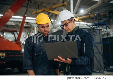 Architect is showing the plan on the laptop. Two male factory workers are in the warehouse 133614728