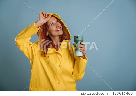 Green thermos in hand, in yellow raincoat. Woman is in the studio against background Green thermos in hand, in yellow raincoat. Woman is in the studio against background 133615406