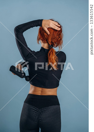 Process of using electric massager, in sportive fitness clothes. Woman is in the studio against background 133615542