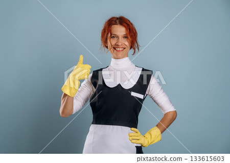Thumb up, showing gesture, maid in uniform. Woman is in the studio against background Thumb up, showing gesture, maid in uniform. Woman is in the studio against background 133615603