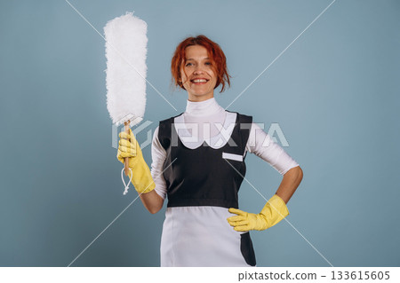 Ready to clean some dust. Maid in uniform. Woman is in the studio against background Ready to clean some dust. Maid in uniform. Woman is in the studio against background 133615605