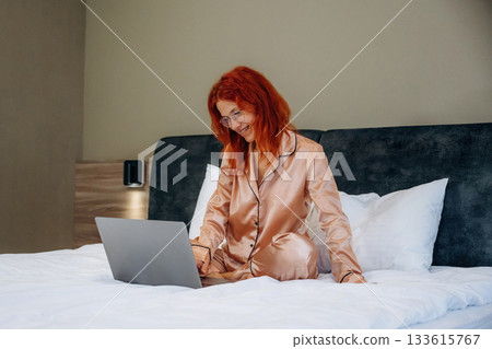 Using laptop, in pajama, on the bed. Woman is in the hotel room, modern interior Using laptop, in pajama, on the bed. Woman is in the hotel room, modern interior 133615767