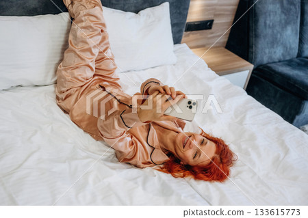 Smartphone in hands, lying down on the bed. Woman is in the hotel room, modern interior Smartphone in hands, lying down on the bed. Woman is in the hotel room, modern interior 133615773