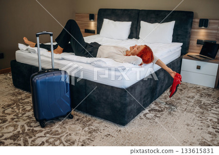 Relaxing, lying down on the bed. Blue suitcase is on the floor. Woman is in the hotel room, modern interior Relaxing, lying down on the bed. Blue suitcase is on the floor. Woman is in the hotel room, modern interior 133615831