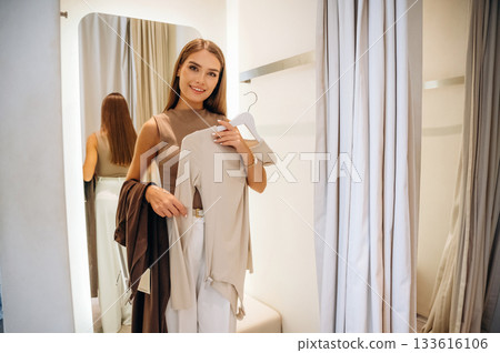 Standing, holding sweater and pants, smiling. A woman tries on clothes in a store, in a fitting room Standing, holding sweater and pants, smiling. A woman tries on clothes in a store, in a fitting room 133616106