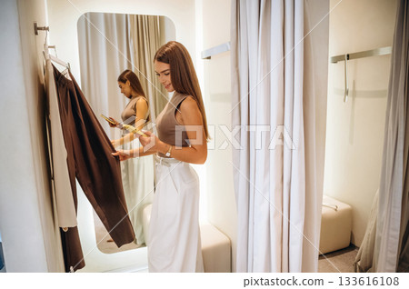 Holding smartphone. A woman tries on clothes in a store, in a fitting room Holding smartphone. A woman tries on clothes in a store, in a fitting room 133616108