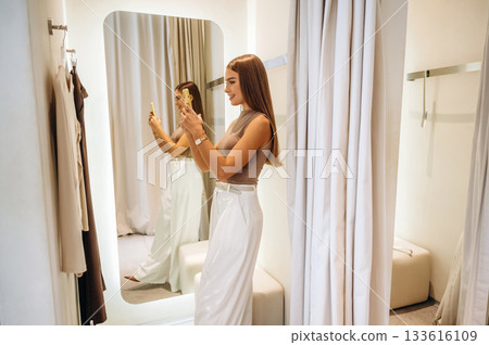 Holding smartphone. A woman tries on clothes in a store, in a fitting room Holding smartphone. A woman tries on clothes in a store, in a fitting room 133616109