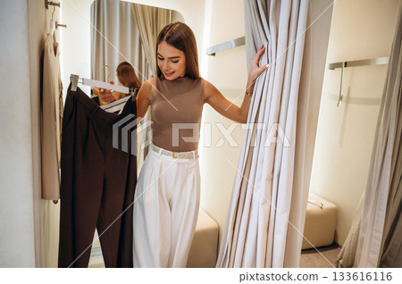 Holding new pants and smiling. A woman tries on clothes in a store, in a fitting room Holding new pants and smiling. A woman tries on clothes in a store, in a fitting room 133616116