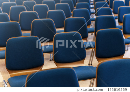 Rows of blue chairs stand in empty hall creating calm geometric pattern. Soft fabric surfaces reflect gentle light forming orderly rhythm and inviting atmosphere 133616269