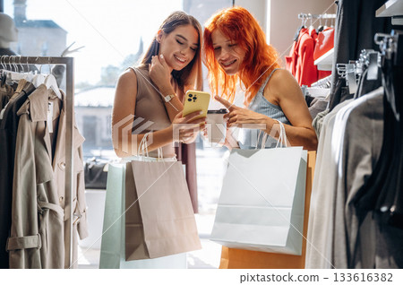 Some information in smartphone, reading. Two female friends are in the retail store, choosing clothes, shopping Some information in smartphone, reading. Two female friends are in the retail store, choosing clothes, shopping 133616382
