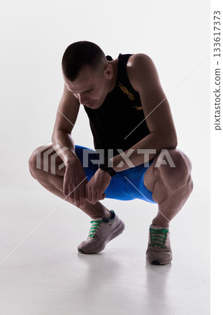 Runner squatting low after training to catch breath in white studio 133617373