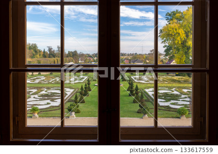 View through Hundisburg palace window reveals ornate garden sculpted hedges and geometric patterns bright sky. Framed perspective creates elegant contrast between interior shadow vivid landscape 133617559