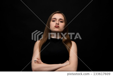 Confident young woman standing with crossed arms in dramatic studio portrait on dark background 133617609