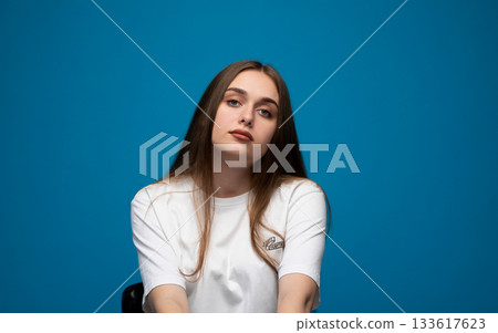 Calm portrait of a young woman with a neutral expression sitting against a clean blue studio background Calm portrait of a young woman with a neutral expression sitting against a clean blue studio background 133617623