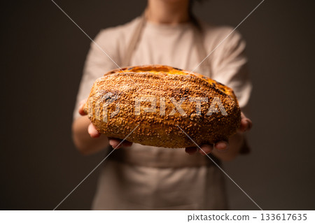 A baker holds out a large crusty seeded loaf, highlighting its texture, sesame seeds and handcrafted artisan quality 133617635