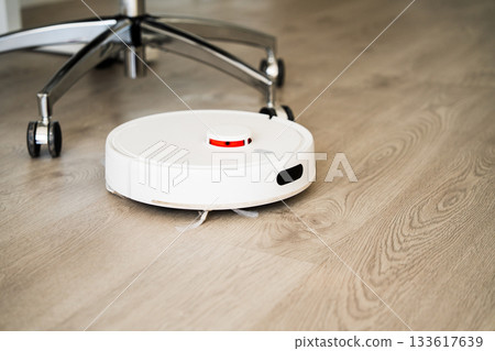 Smart robot vacuum cleaner moving across wooden floor near office chair for efficient automated cleaning 133617639