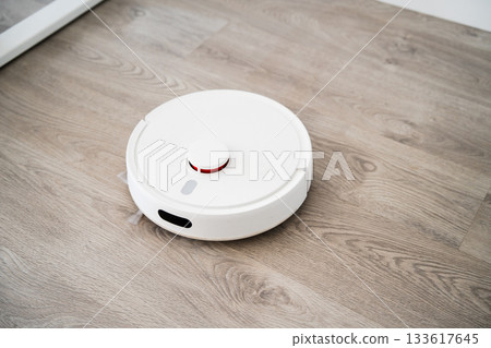 White robotic vacuum positioned on wood flooring while cleaning open interior zone in bright modern household 133617645