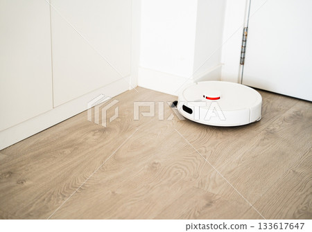 Smart robotic vacuum cleaning wooden floor near white cabinet in bright minimalist home interior environment Smart robotic vacuum cleaning wooden floor near white cabinet in bright minimalist home interior environment 133617647