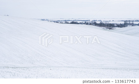 Winter rural landscape with snowy fields and distant village under cloudy sky 133617743