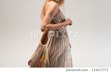 Side view of woman in light striped dress with eco mesh bag and baguette isolated on gray background Side view of woman in light striped dress with eco mesh bag and baguette isolated on gray background 133617773