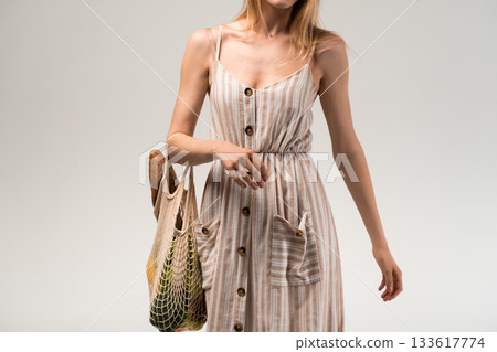 Woman in striped summer dress holding eco mesh bag with bread and vegetables on neutral background 133617774