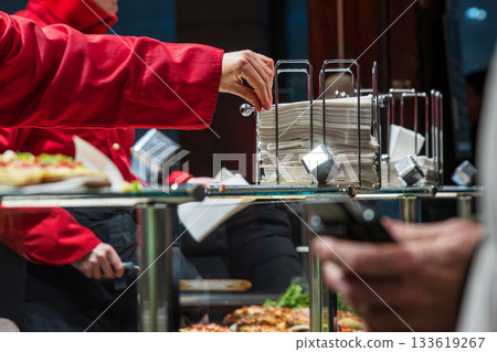 Vendor in a red jacket pulling napkins from a metal dispenser, serving food trays for customers in front. Street food culture and busy market atmosphere. 133619267