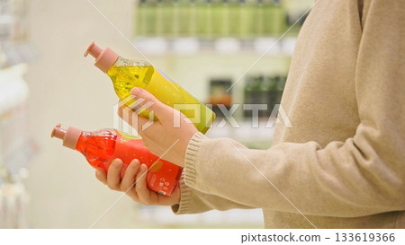 Woman comparing two shampoo bottles in a store aisle, carefully reading labels and ingredients for the best haircare and cosmetic choice. 133619366