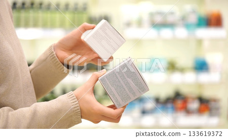 Female shopper reading ingredient labels, comparing and choosing a cosmetic product or cream, focusing on packaging details in a beauty store. The concept of conscious choice 133619372