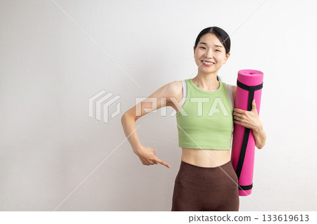 一位微笑的女子手持瑜珈墊，指著自己的腰部。 133619613