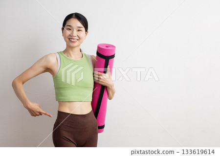 一位微笑的女子手持瑜珈墊，指著自己的腰部。 133619614