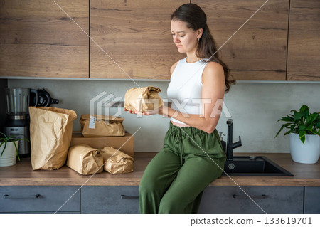 Young woman holding a brown paper bag with meal kit ingredients in a modern kitchen. Concept of convenient recipe-based grocery delivery and mindful home cooking. 133619701