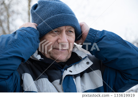 Senior man wearing blue winter knitted hat and jacket adjusts his hat outdoors. Man preparing for cold weather. Winter season. Outdoors. 133619919