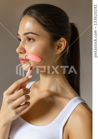 Young woman using a pink facial roller on her cheek, demonstrating a relaxing skincare massage routine on a soft studio background. Young woman using a pink facial roller on her cheek, demonstrating a relaxing skincare massage routine on a soft studio background. 133620292