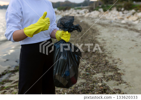 Volunteer collecting beach litter into a black trash bag during coastal cleanup, highlighting pollution, waste problem, environmental protection and eco awareness. 133620333