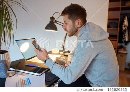 Man sitting at desk with laptop and holding smartphone during remote work 133620335