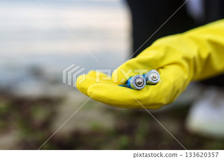 Close-up of a volunteer wearing yellow gloves holding discarded batteries collected on a polluted beach. Environmental cleanup, toxic waste, coastline protection and eco awareness. 133620357